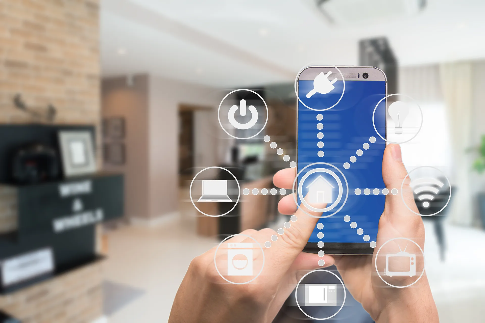 A person uses a smartphone with a smart home app interface to control various connected appliances and devices like lights, a laptop, and a washing machine.